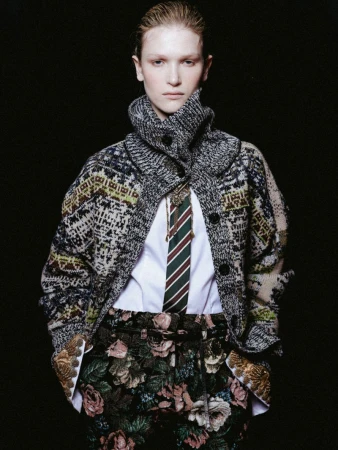 Fashion editorial shot of a young adult woman in a patterned coat and floral pants against a stark black backdrop in a studio setting.