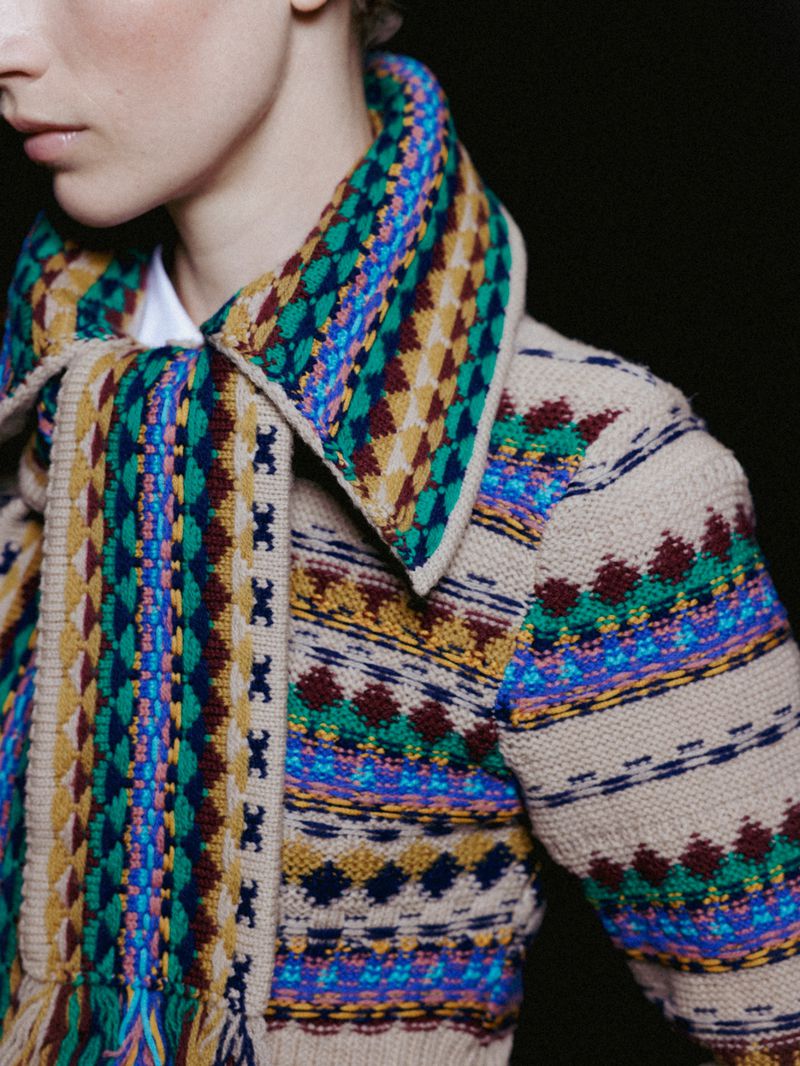 Close-up shot of a fair-skinned person with a patterned tan cardigan with blue, yellow, and green on a dark background.