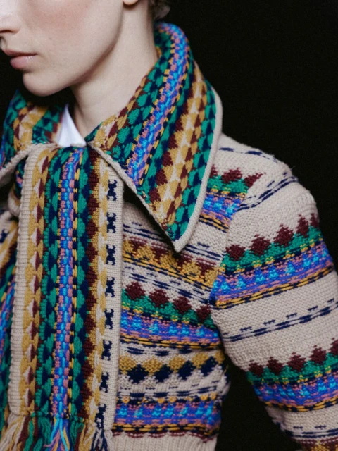 Close-up shot of a fair-skinned person with a patterned tan cardigan with blue, yellow, and green on a dark background.