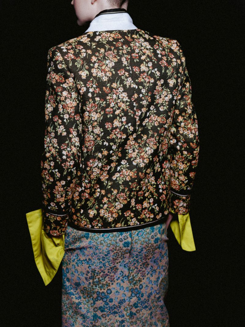 Back view of a person wearing a dark floral jacket and a floral skirt with long yellow shirt sleeves, posing in a dark studio.