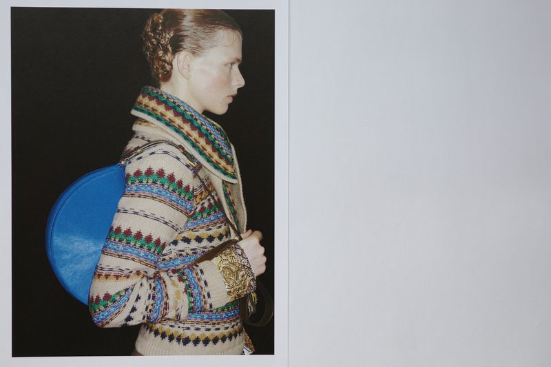 Profile portrait of an adult woman wearing a patterned jacket and a blue round handbag, set against a black background.