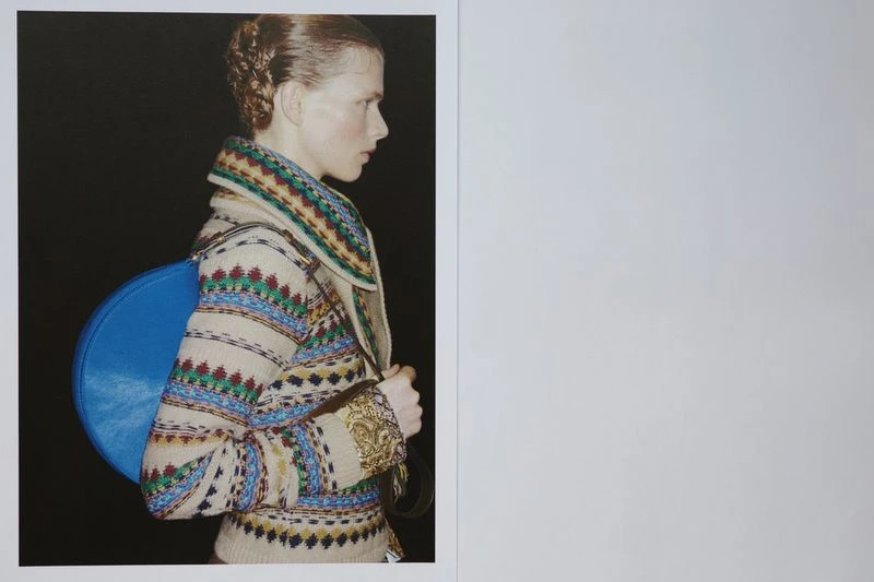 Profile portrait of an adult woman wearing a patterned jacket and a blue round handbag, set against a black background.