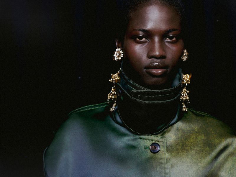 Close-up portrait of a dark-skinned woman with jewelry and a green top, set against a dark background in studio lighting.