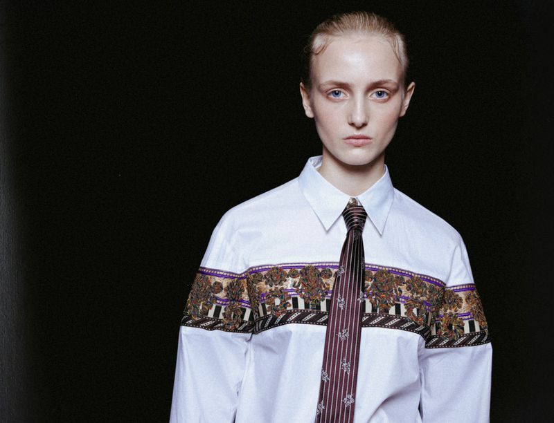 Fashion portrait of a young woman with blue eyes wearing a white shirt with patterned detail and a dark tie against a black backdrop.