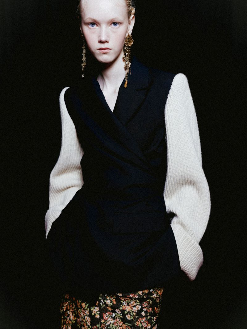 A fair-skinned person with long earrings wearing a black jacket, floral skirt, and white sleeves poses against a dark backdrop.