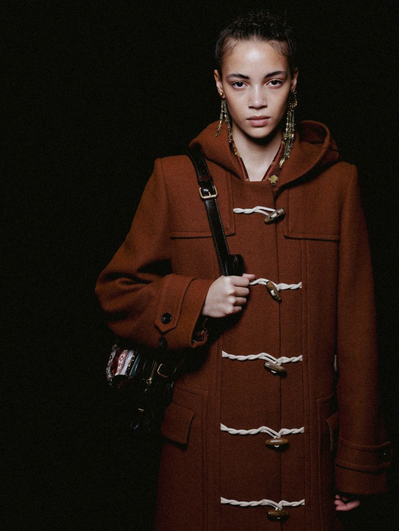 A woman poses in a brown toggle coat with a shoulder bag against a dark backdrop, showcasing fashion and style in a studio setting.