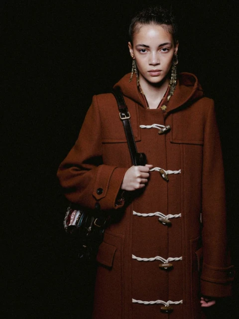 A woman poses in a brown toggle coat with a shoulder bag against a dark backdrop, showcasing fashion and style in a studio setting.