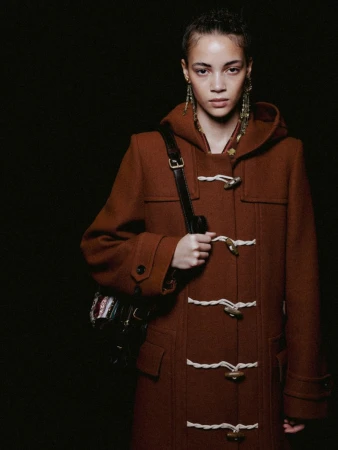 A woman poses in a brown toggle coat with a shoulder bag against a dark backdrop, showcasing fashion and style in a studio setting.
