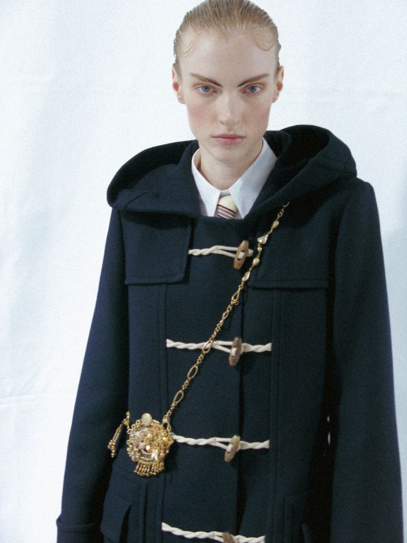 A fair-skinned model wears a dark blue duffel coat with toggle closures and a gold chain embellishment in a fashion-style, eye-level shot.