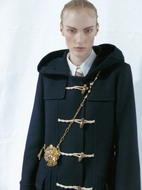 A fair-skinned model wears a dark blue duffel coat with toggle closures and a gold chain embellishment in a fashion-style, eye-level shot.
