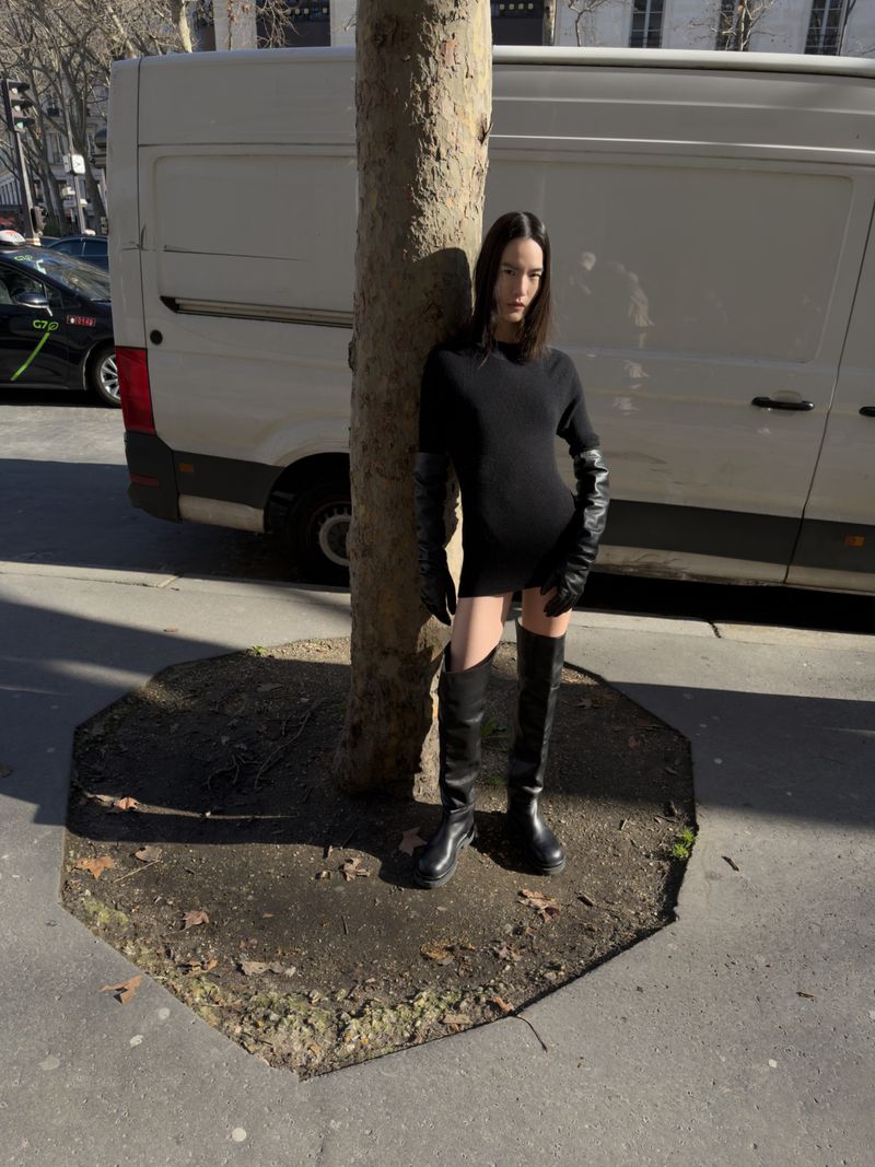 Woman in a black dress and long gloves stands next to a tree in a city street with a white van and cars in the background.