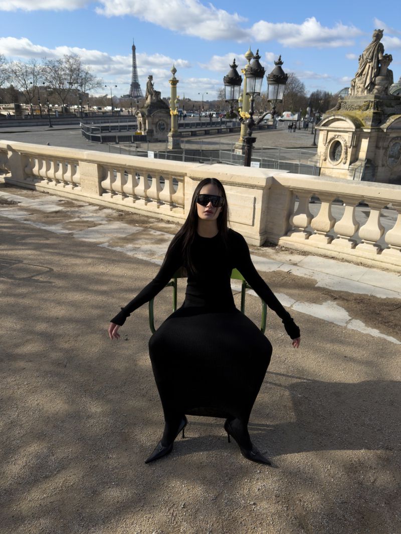 Woman in black dress seated outdoors in Paris with Eiffel Tower in background on a sunny day with blue sky and few clouds.