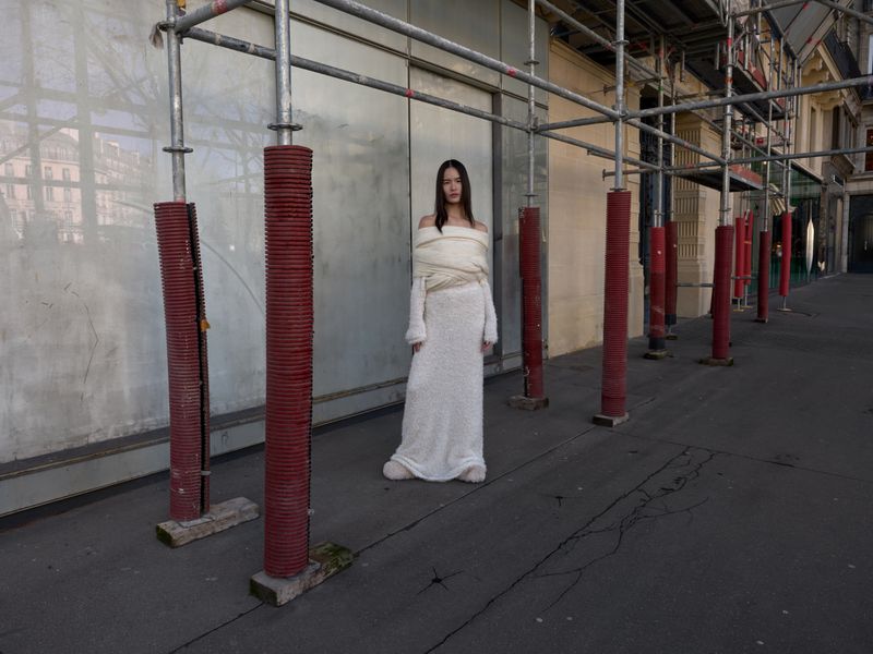 Fashion shot of a young woman in a white dress posing between metal construction scaffolding on an urban street during daytime.
