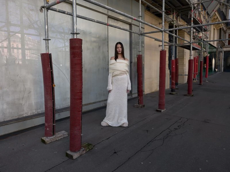 Fashion shot of a young woman in a white dress posing between metal construction scaffolding on an urban street during daytime.
