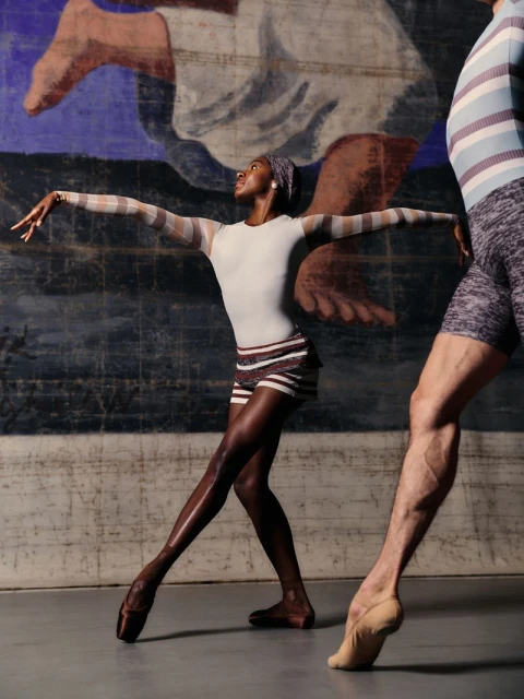 Two dancers in ballet attire pose in front of an aged mural featuring painted figures and a blue and gray textured background.