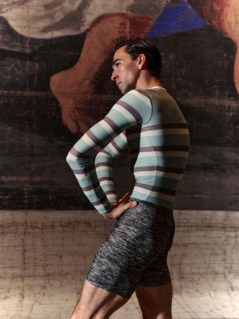 Adult male posing in striped long sleeve shirt and gray athletic shorts in front of an abstract mural in a studio setting. 