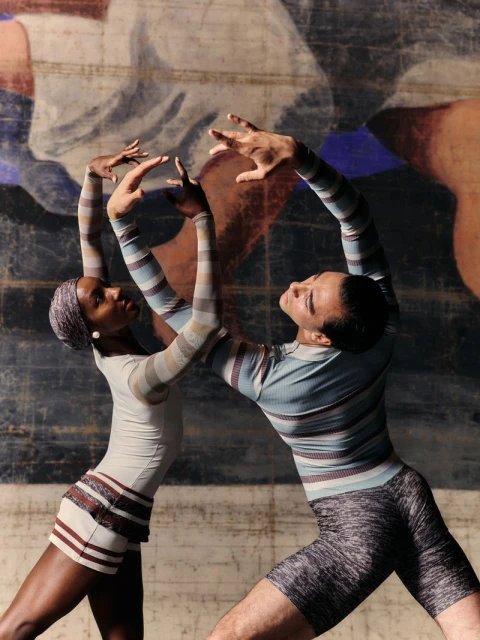 Two dancers in a studio, woman in striped top and mini-skirt, man in striped top and shorts, dancing in front of an abstract mural.