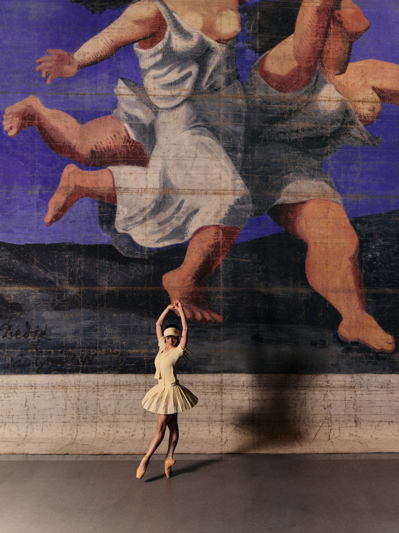 A ballerina in a yellow tutu poses en pointe in front of a painted backdrop featuring classical figures in white dresses.