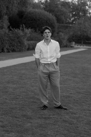 Monochrome shot of a young adult man in a white shirt and pleated trousers, standing on a lawn in a park-like setting with trees and a path.