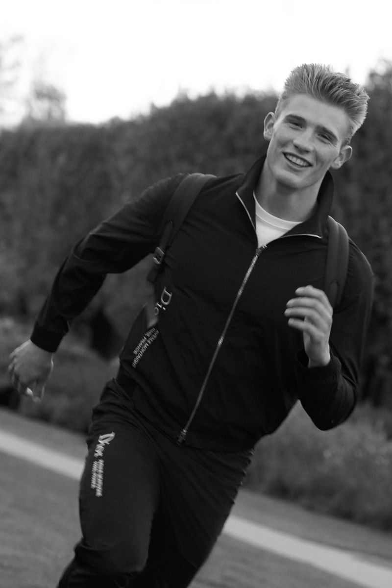 Athletic blond man in dark athletic suit runs towards the camera outdoors with a green background, captured in black and white.