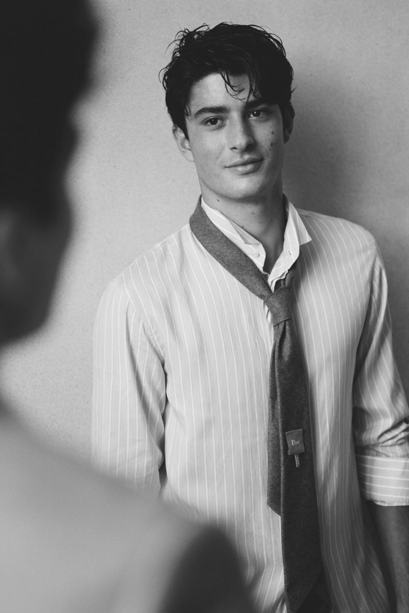 Monochrome portrait of a young adult male wearing a striped shirt and loosened tie, with a blurred figure in the foreground.