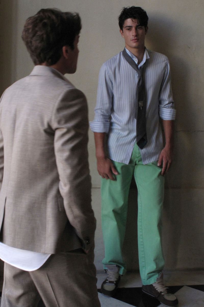 Two young adult men pose in a fashion shot, one wearing a light brown suit, the other in an unbuttoned striped shirt, tie, and bright green pants.