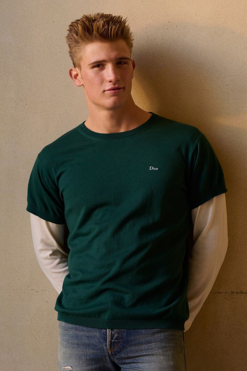 A fair-skinned adult male is posed indoors, wearing a layered green t-shirt and a long-sleeved shirt, with faded denim jeans. 