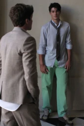 Model in a striped shirt, loose tie, and green trousers leaning against a wall facing a man in a beige tailored suit.