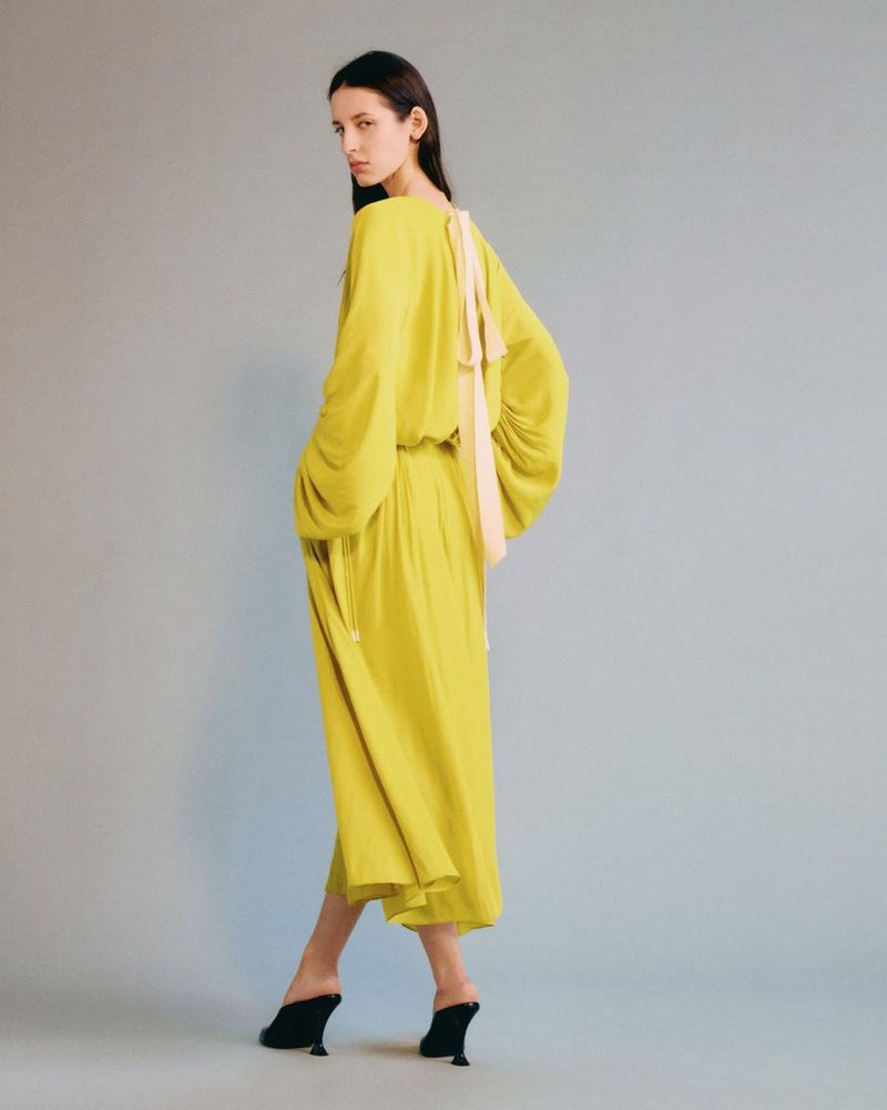 A slender woman is captured in a studio setting, showcasing a chartreuse dress with a light pink bow accent and black heeled shoes.