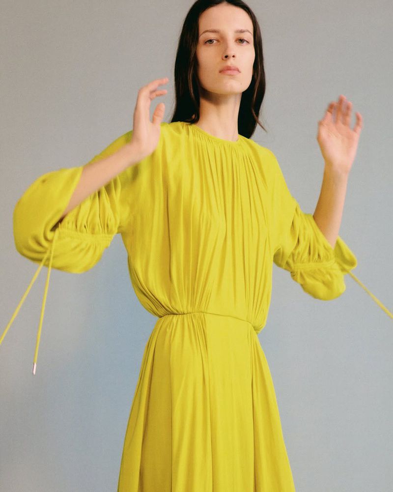 A woman poses in a studio wearing a yellow-green dress with long sleeves, her hands raised against a neutral grey background in a fashion shot.