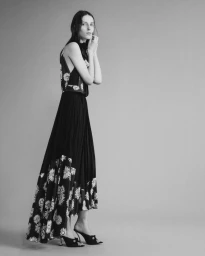 Model Valery Sergeeva poses in profile wearing a black floral pleated maxi dress and kitten heel mules against a studio backdrop.