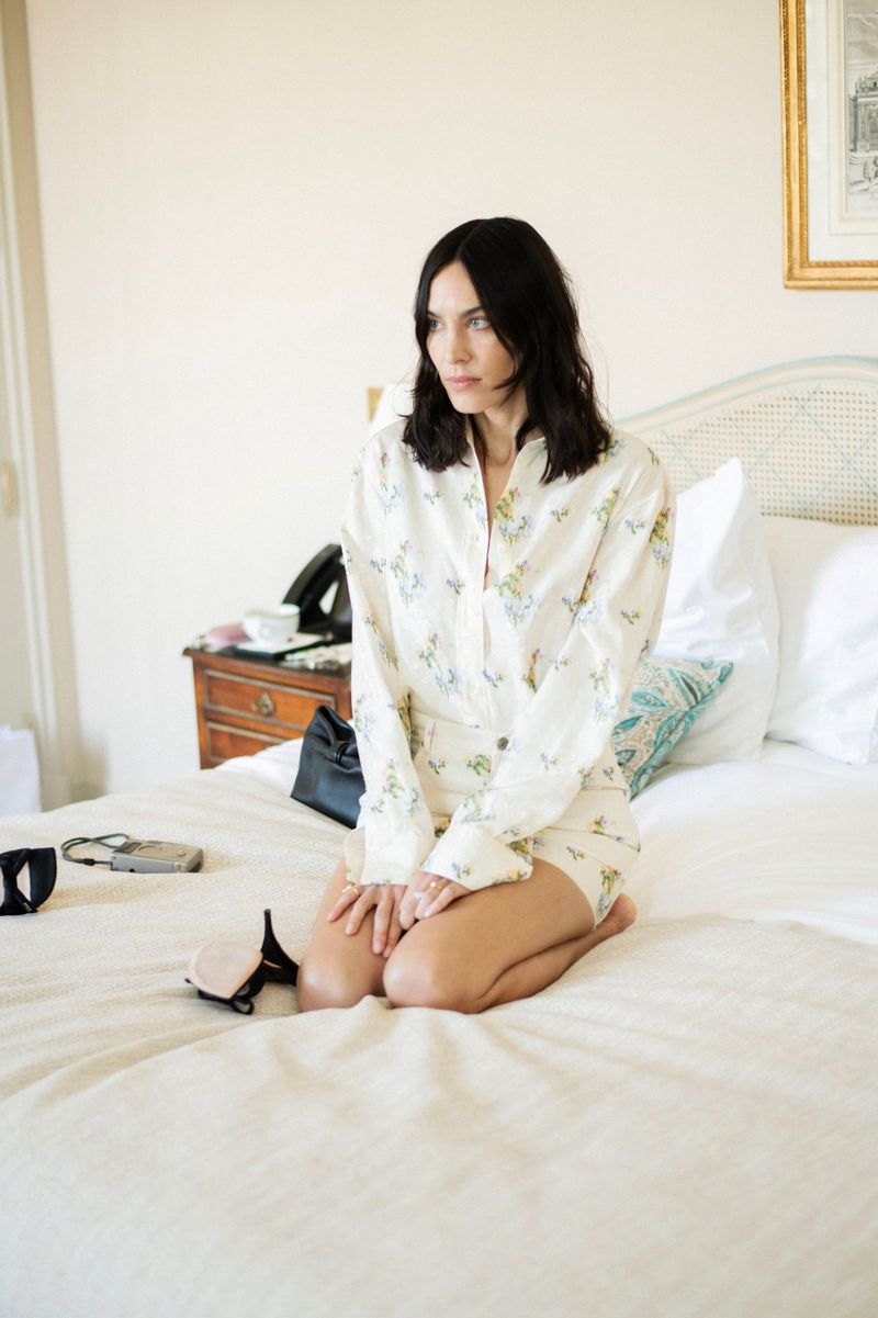 Woman in floral pajamas sitting on a bed with white bedding, bedside table with phone, and framed art in the background.