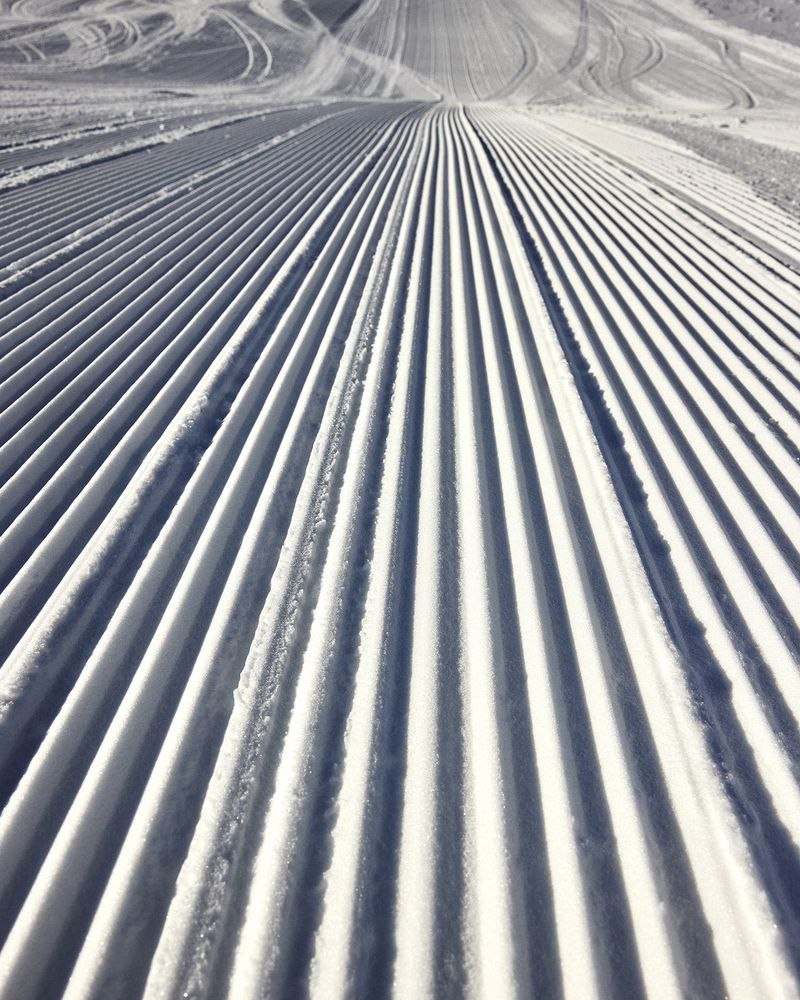 Freshly groomed ski slope with corduroy pattern on a sunny day, creating a visually appealing texture in a winter mountain setting.