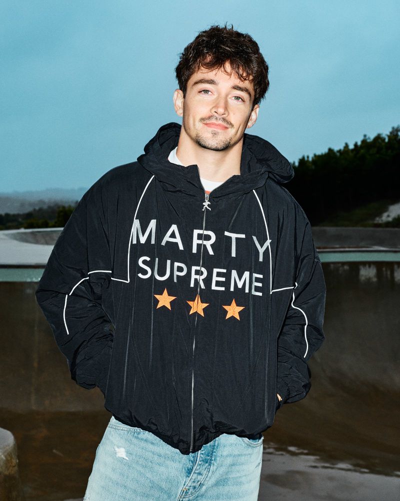 Man in black Marty Supreme hooded jacket poses outdoors. He wears jeans and has a slight smile in a medium shot.