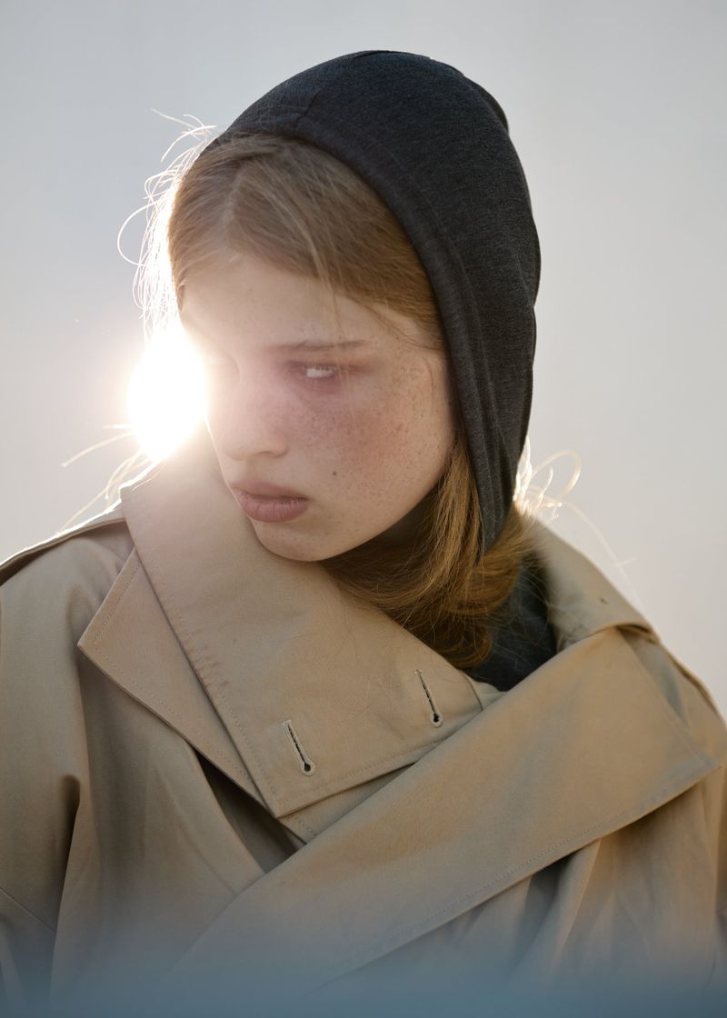 Close-up portrait of a person with freckles wearing a dark grey hoodie and a tan trench coat, captured in backlit, hazy sunlight.