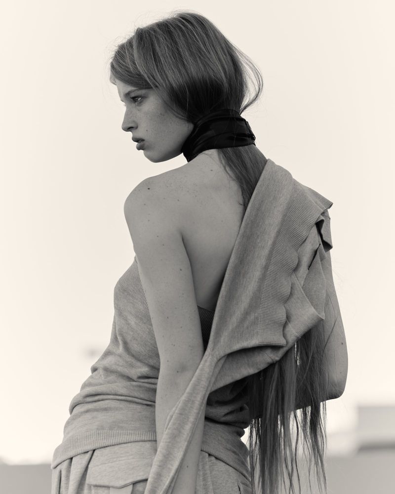 Monochromatic portrait of a fair-skinned young woman with long hair, wearing a grey sweater and black neck scarf, looking to the side.