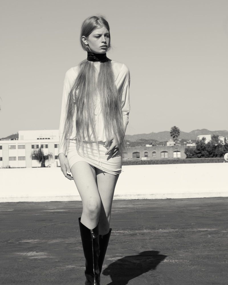 A woman poses in a short dress and boots outdoors on a sunny day with a cityscape and mountains in the background in a black and white image.