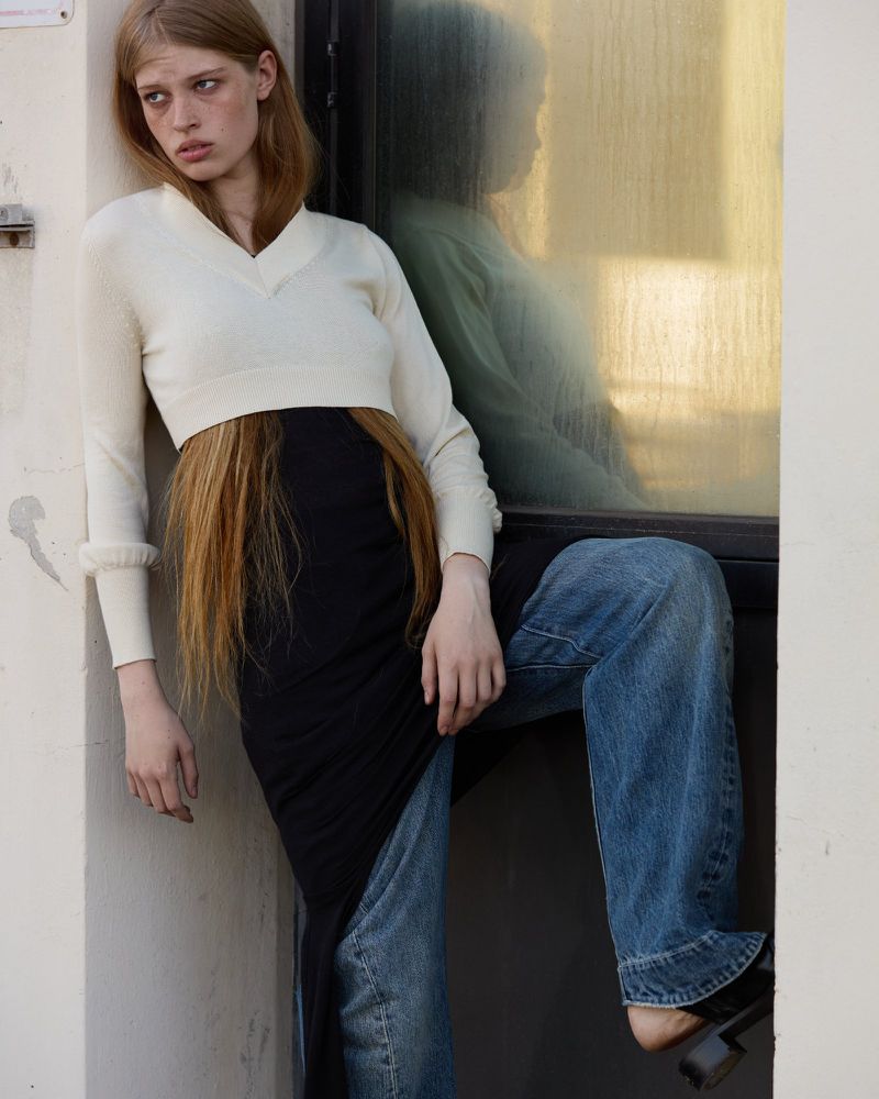 A young fair-skinned woman poses by a sunny window in a cream sweater, black dress and jeans with chunky heeled shoes in urban setting.