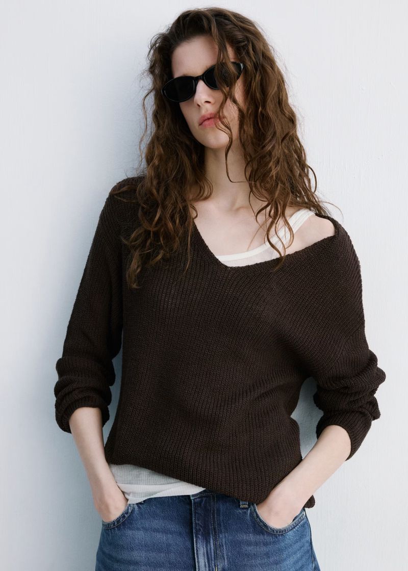 A woman in sunglasses and a dark brown sweater stands against a white wall, wearing jeans and posing with her hands in her pockets.