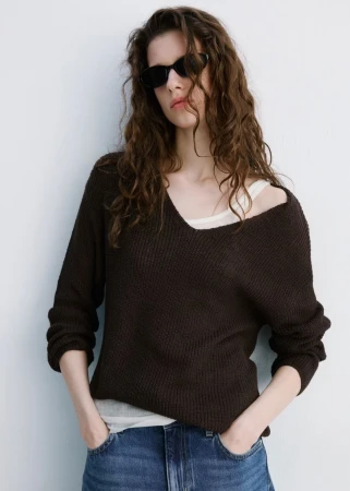 A woman in sunglasses and a dark brown sweater stands against a white wall, wearing jeans and posing with her hands in her pockets.