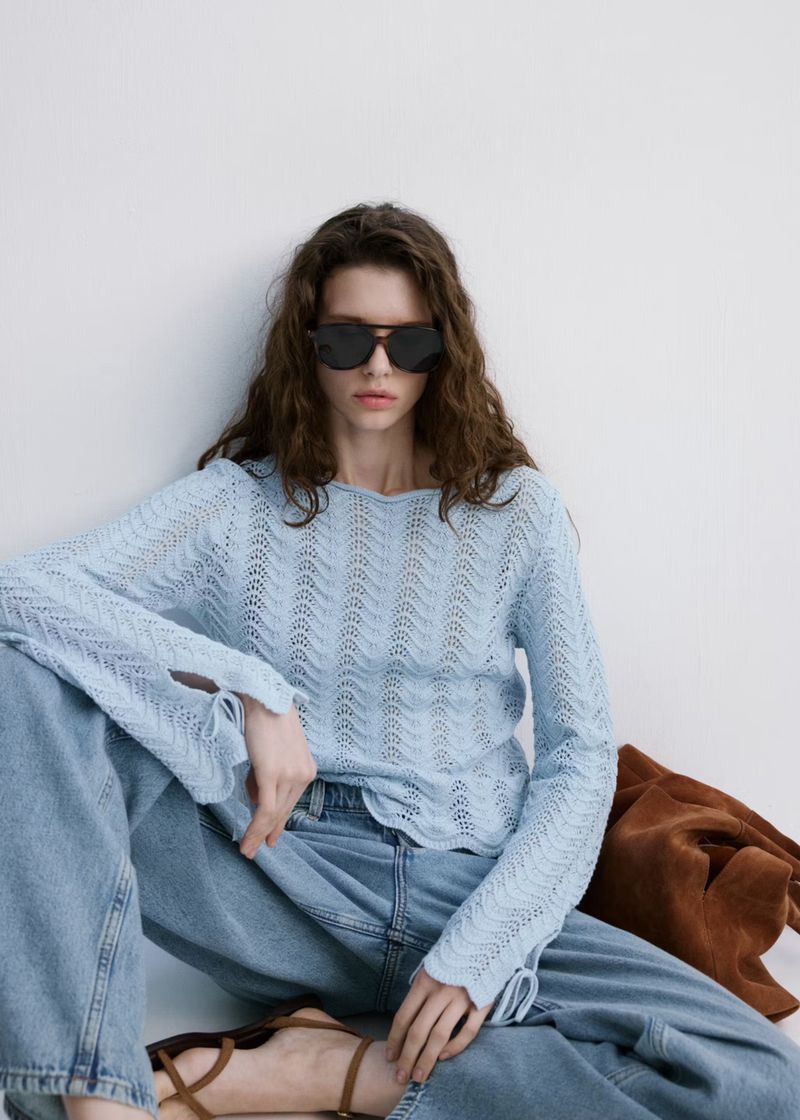 A woman wearing sunglasses, light blue knitted shirt, and denim jeans is sitting indoors with a brown bag on a white background.