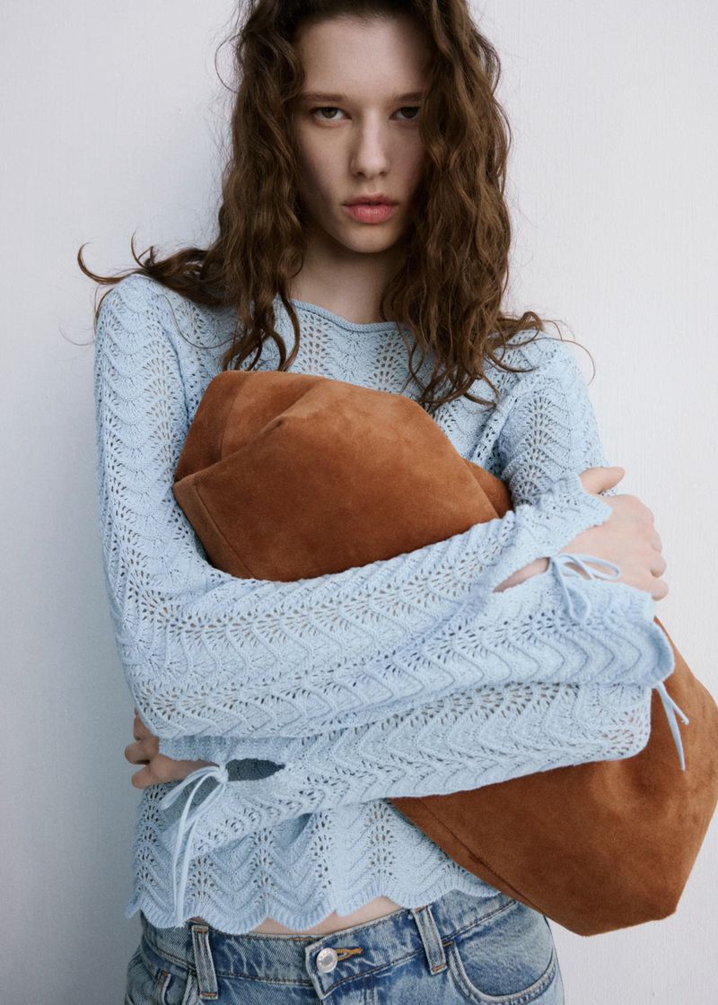 A young woman with curly brown hair poses in a light blue, long-sleeve top and denim jeans, holding a suede tote bag indoors.