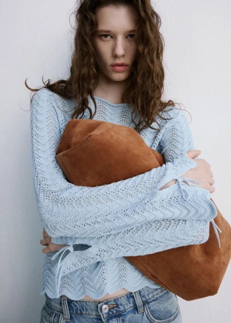 A young woman with curly brown hair poses in a light blue, long-sleeve top and denim jeans, holding a suede tote bag indoors.
