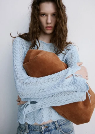 A young woman with curly brown hair poses in a light blue, long-sleeve top and denim jeans, holding a suede tote bag indoors.