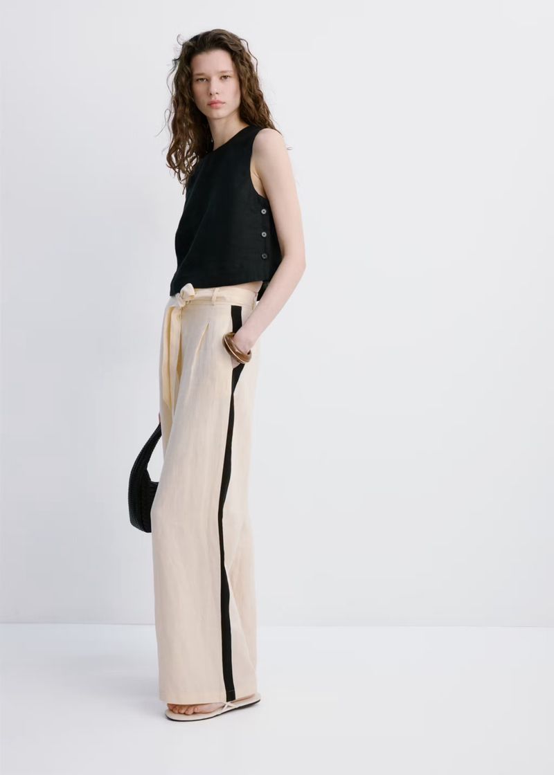 Woman in a black sleeveless top and beige pants with a black stripe posing in a white studio. She carries a black bag.