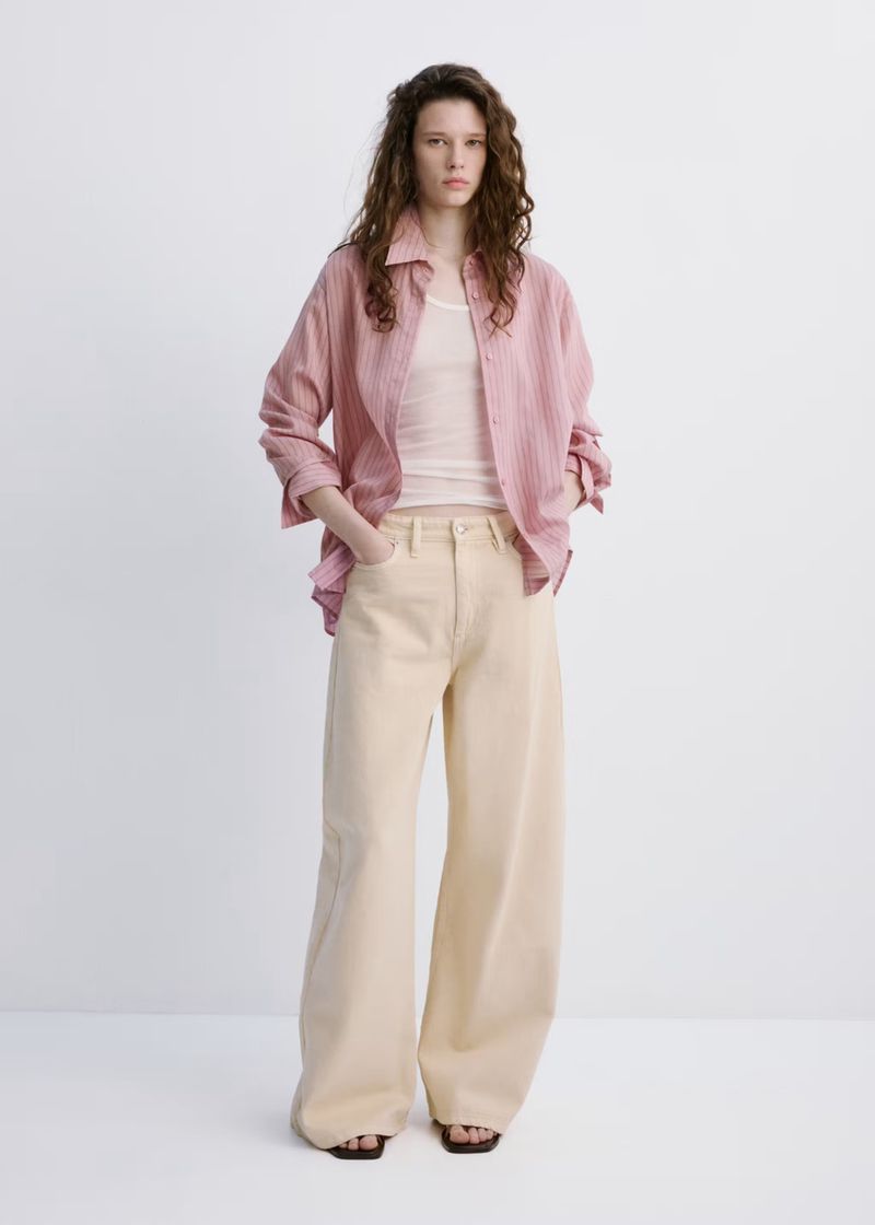 Young woman in a pink striped shirt, white top, and beige wide-leg jeans poses in a bright photo studio for a fashion editorial.