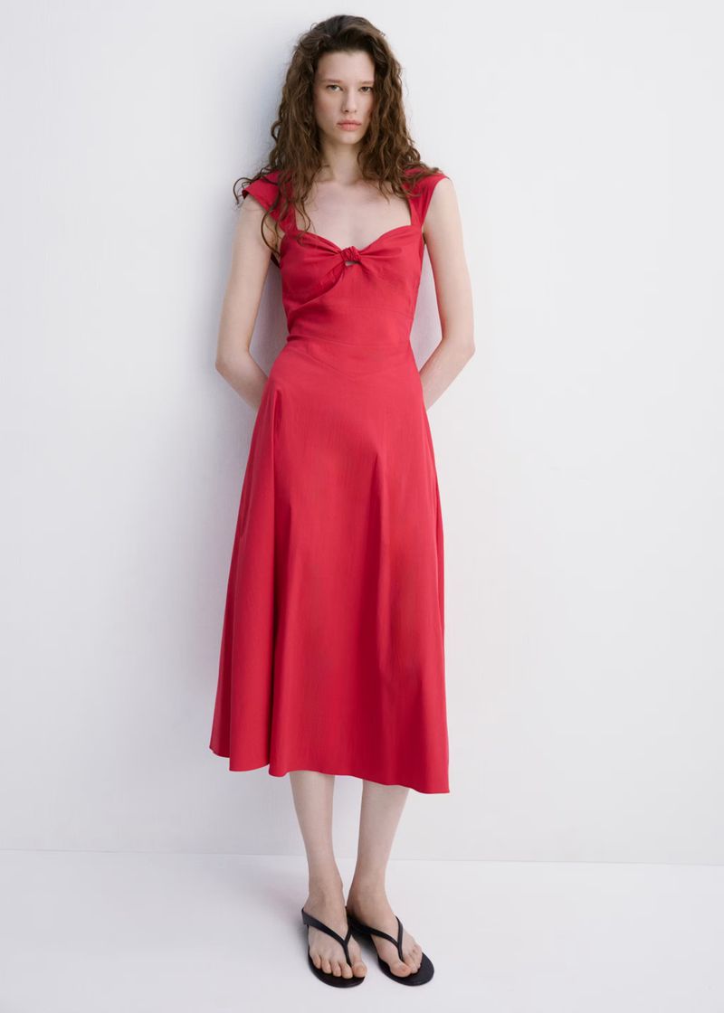 A slender woman with curly brown hair poses in a studio wearing a red midi dress and black flip-flops against a white backdrop.
