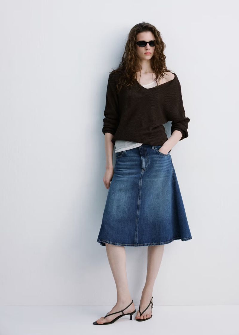 Full-length studio shot of a woman in a brown V-neck sweater, blue denim skirt, and black heeled sandals against a white wall.