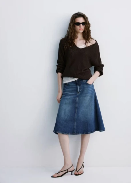 Full-length studio shot of a woman in a brown V-neck sweater, blue denim skirt, and black heeled sandals against a white wall.
