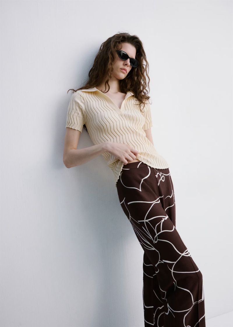Adult female fashion model with brown curly hair poses in polo shirt and patterned trousers against a white wall in soft daylight.
